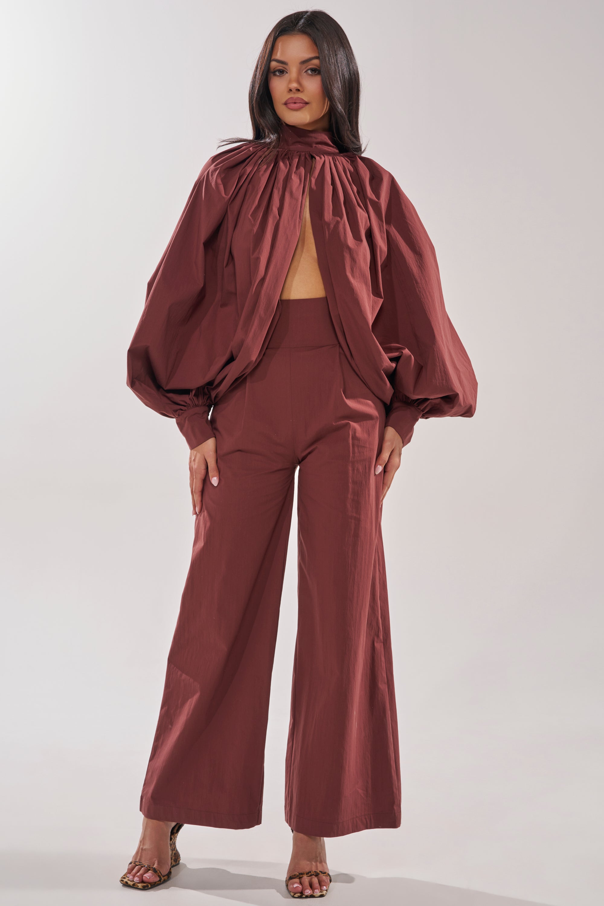 A woman wears the GOLD COAST WIDE LEG PALAZZO PANT IN BROWN, styled with a matching long-sleeve, high-neck top featuring a front cutout. She stands with her hands at her sides against a plain light background. 1