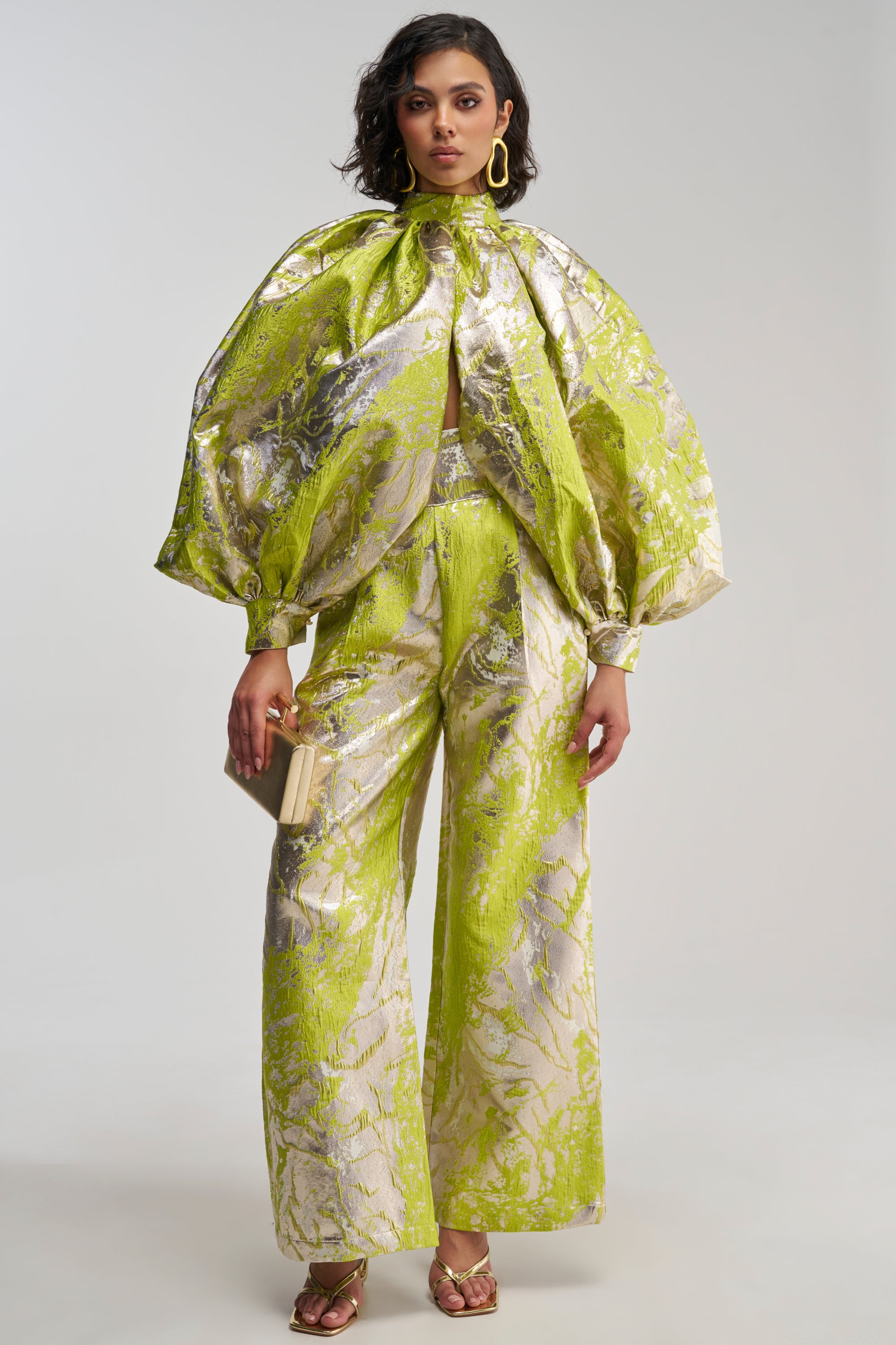 Dressed for a stylish holiday party, the woman pairs shimmering gold heels and statement earrings with the ELECTRIC GARDEN BROCADE PALAZZO PANT—featuring a striking green and silver metallic pattern in a wide-leg silhouette—and a matching puff-sleeve blouse. She completes her look with a chic small clutch.