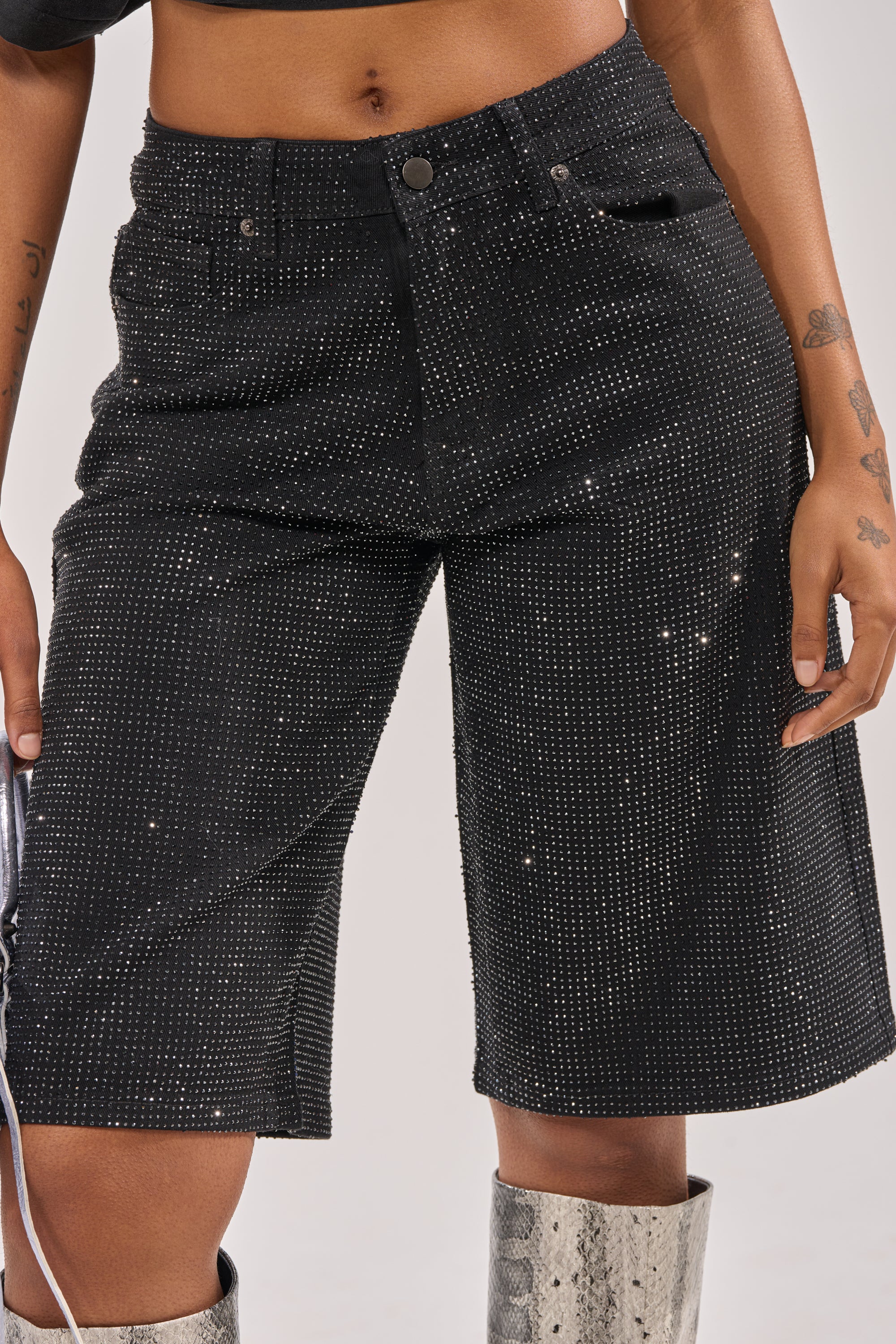 A person wearing the BLING BLING RHINESTONE BERMUDA DENIM SHORT IN BLACK, a cropped top, and silver snakeskin boots. Their visible hands reveal small tattoos on their arms and fingers. 2
