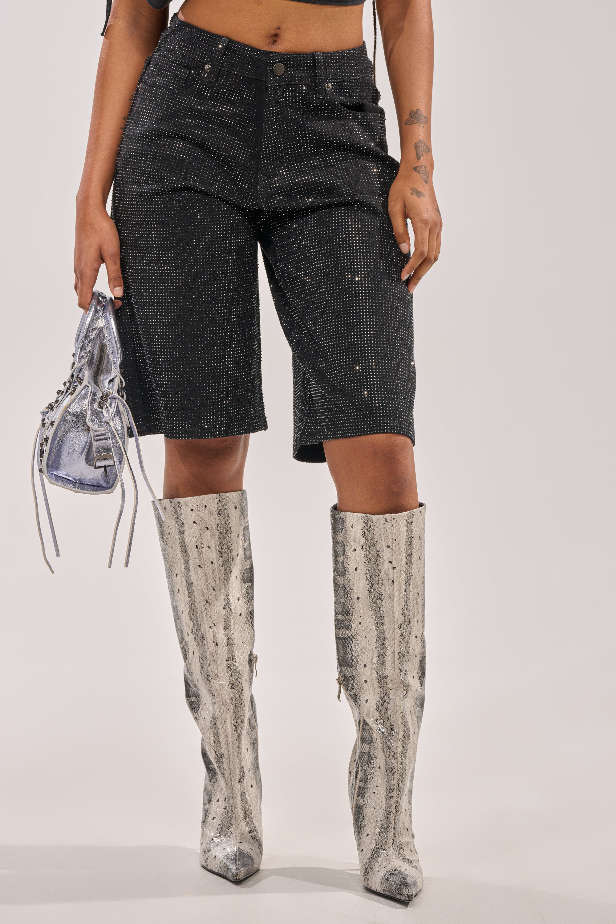 A person wears the BLING BLING RHINESTONE BERMUDA DENIM SHORT IN BLACK, paired with a metallic silver purse and knee-high silver snakeskin boots, standing against a plain background. 3