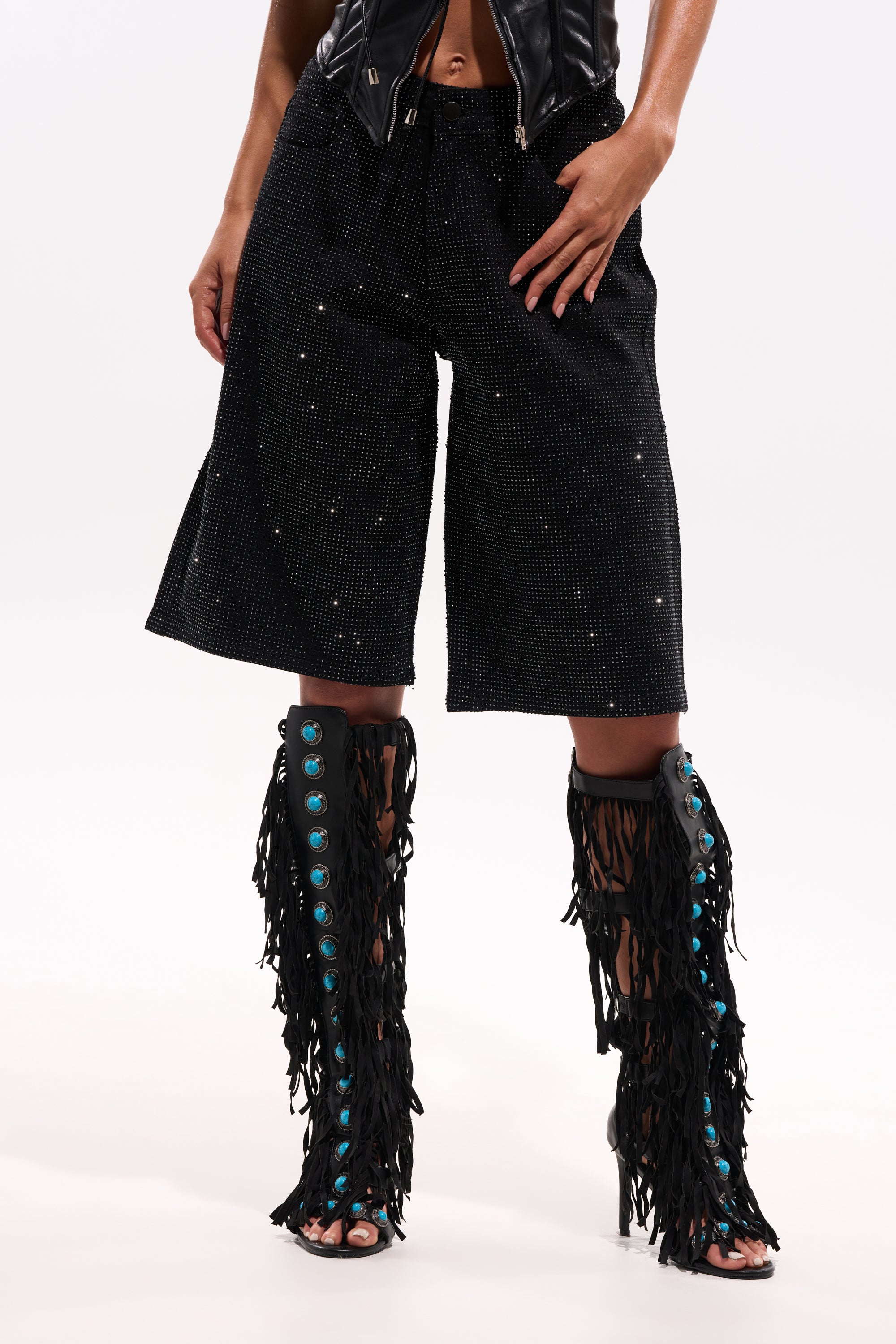 A person wearing the BLING BLING RHINESTONE BERMUDA DENIM SHORT IN BLACK and black fringe knee-high open-toe boots with turquoise button details, standing against a plain white background. 1