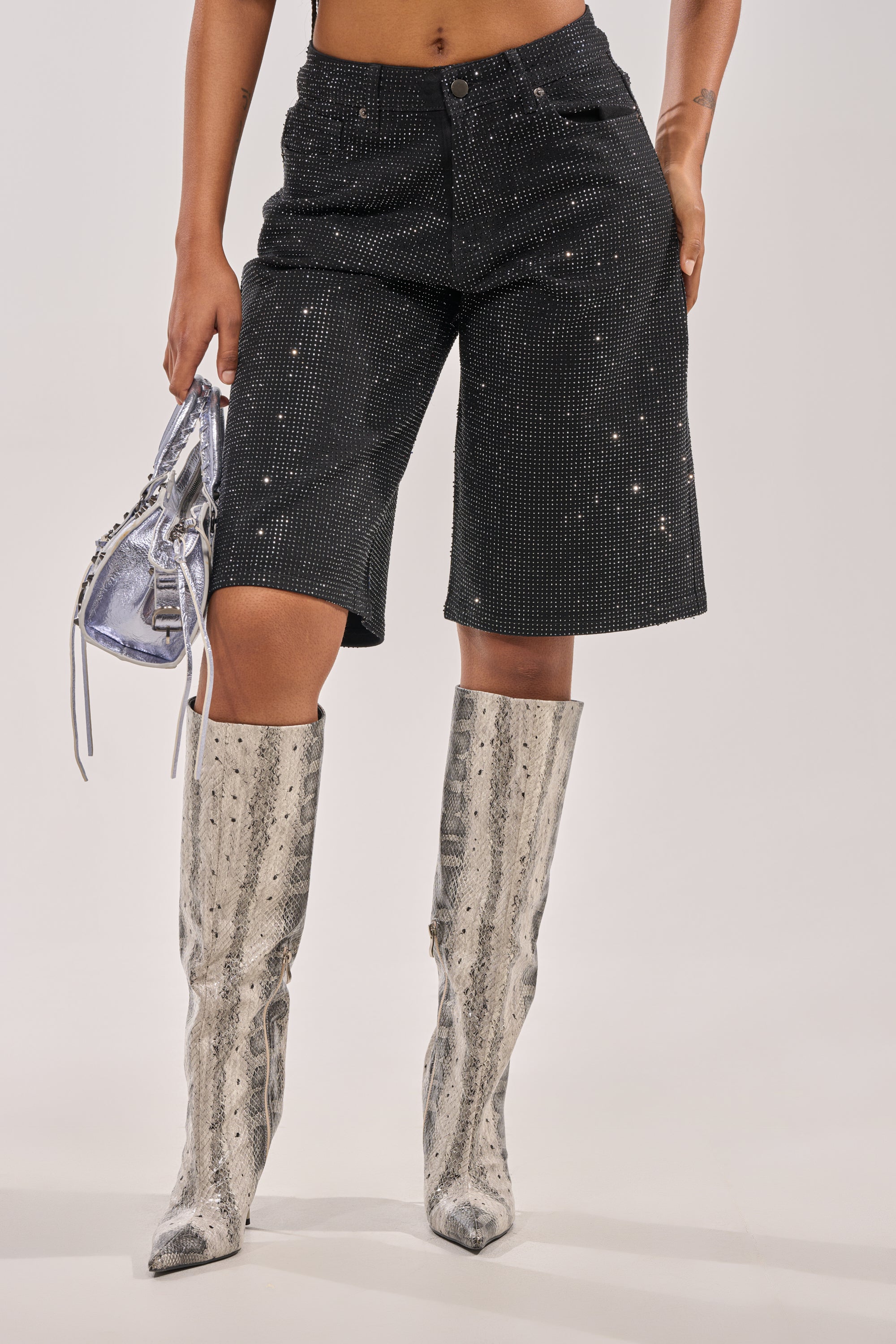 Someone wears the BLING BLING RHINESTONE BERMUDA DENIM SHORT IN BLACK with rhinestone details and knee-high silver snakeskin boots, holding a small metallic silver handbag against a plain background. 4