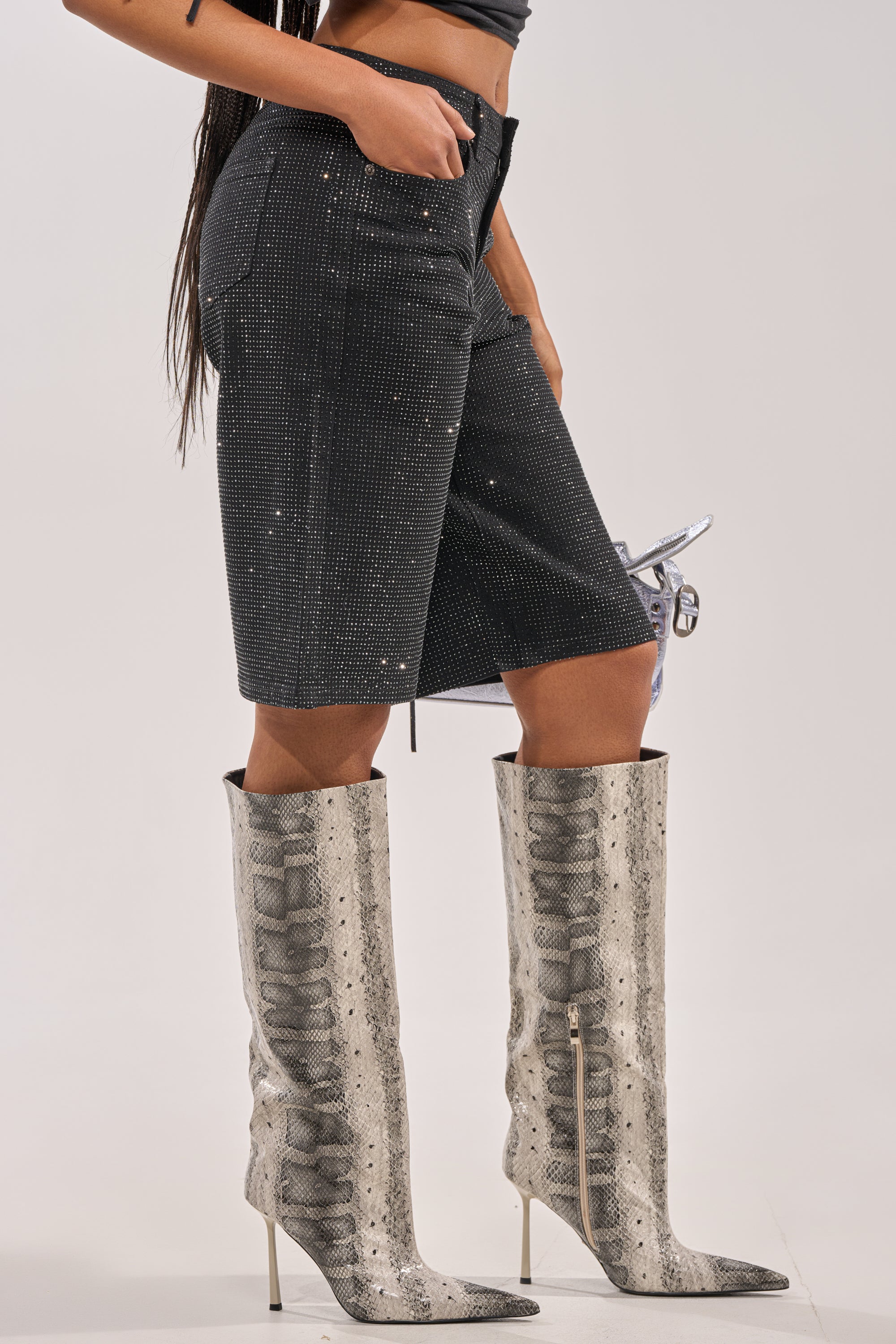 A person wears BLING BLING RHINESTONE BERMUDA DENIM SHORT IN BLACK with rhinestone embellishments, a black crop top, tall gray snakeskin-patterned pointed-toe high-heeled boots, and holds a small shiny silver handbag. 5