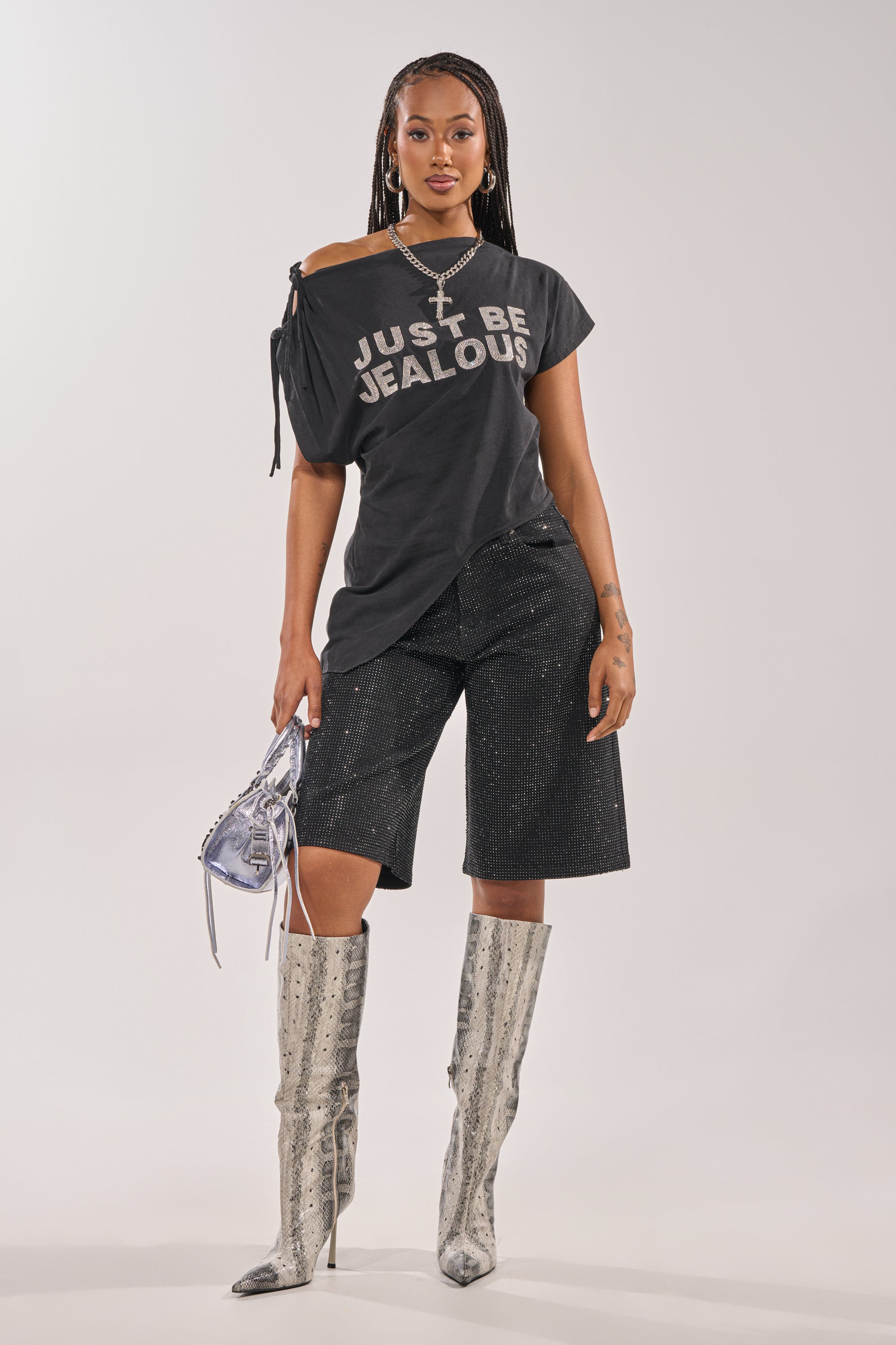 A woman stands confidently in a black "JUST BE JEALOUS" tee, BLING BLING RHINESTONE BERMUDA DENIM SHORT IN BLACK, silver knee-high snakeskin boots, layered necklaces, and a metallic silver handbag. 7