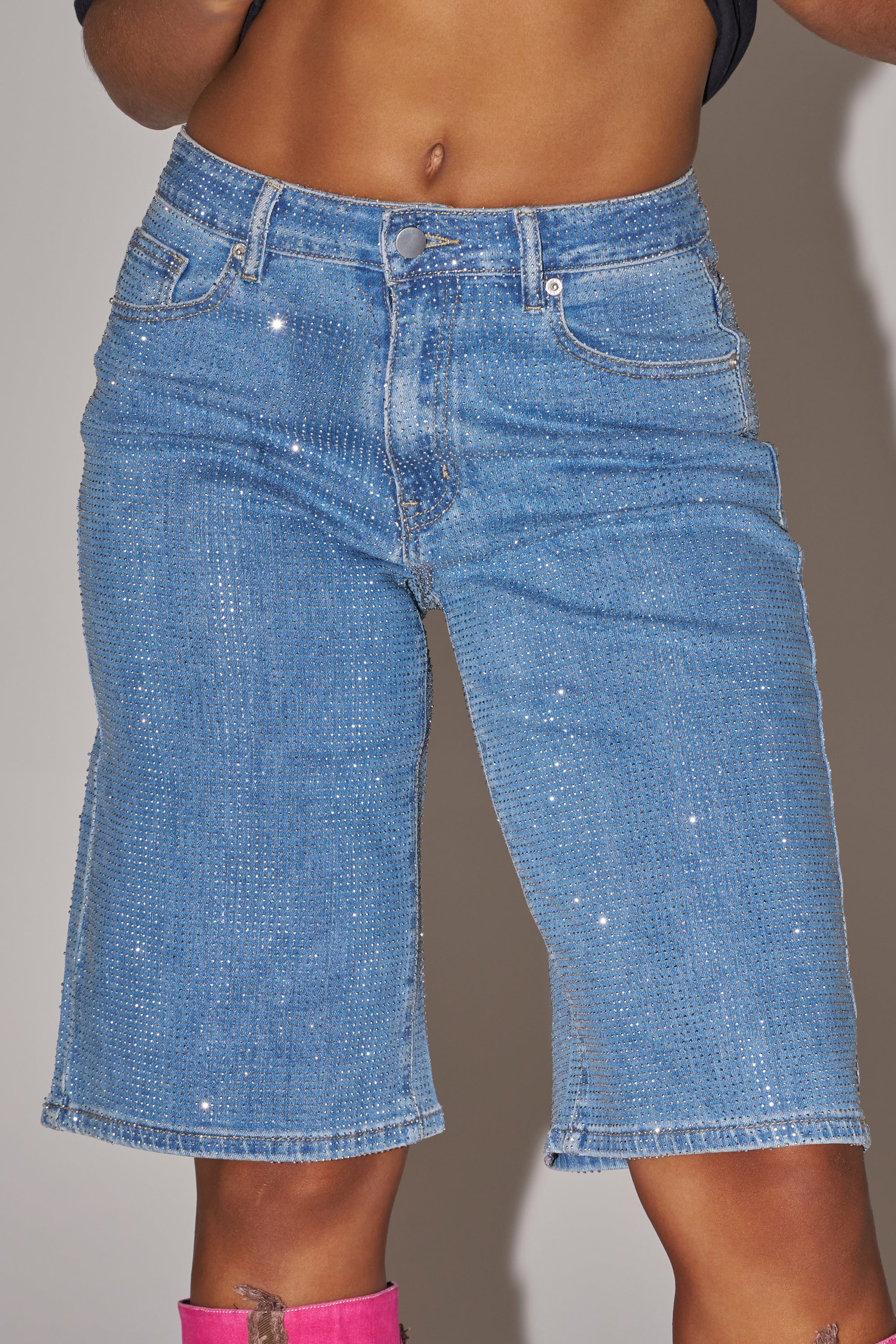 A close-up shot shows only the torso and upper legs of a person wearing the BLING BLING RHINESTONE DARK BLUE DENIM BERMUDA SHORT with sparkling embellishments, paired with a dark crop top that reveals the midriff. 10