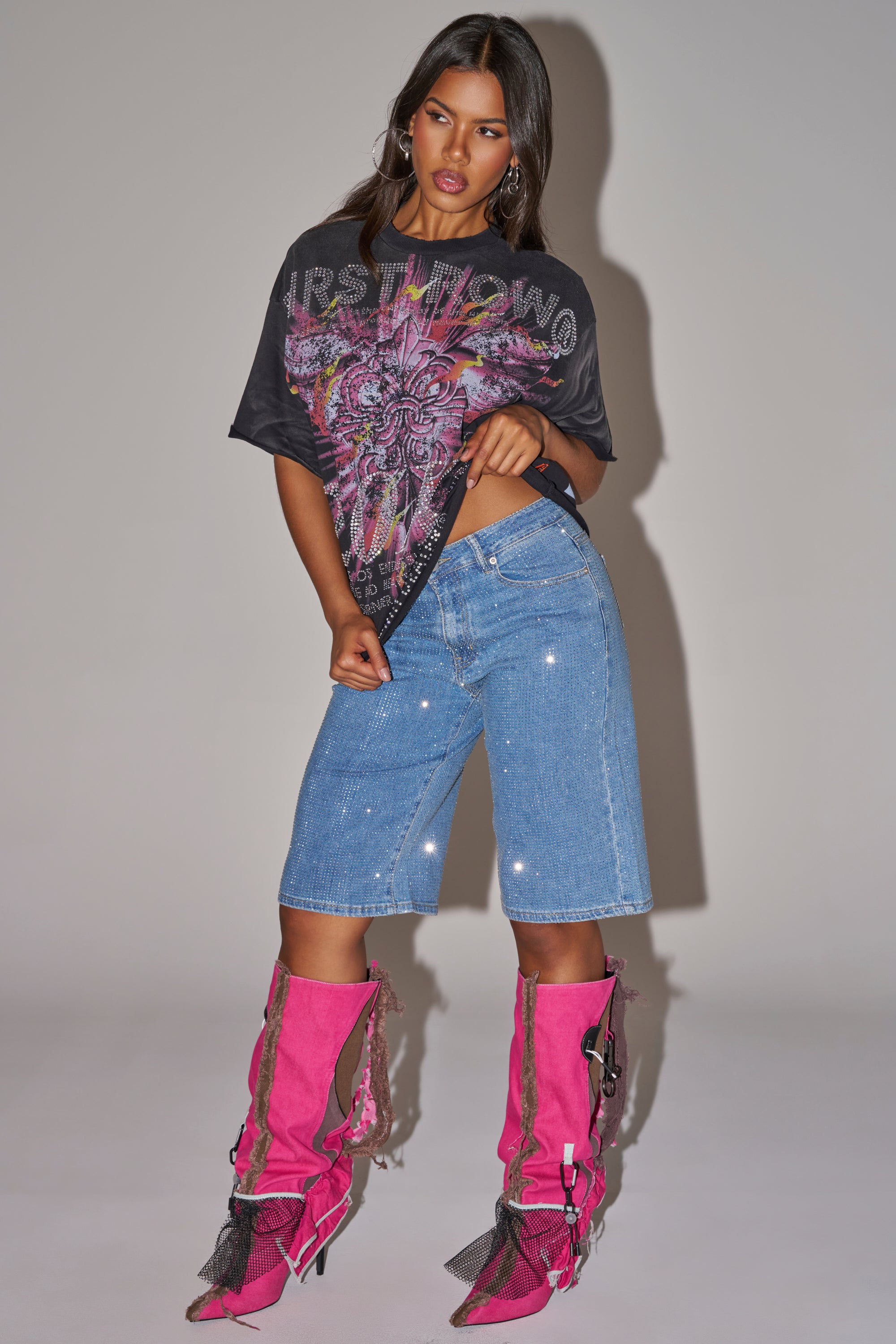 A woman poses in a graphic tee, BLING BLING RHINESTONE DARK BLUE DENIM BERMUDA SHORT, and tall bright pink boots with fringe, buckles, and tassels against a plain light background. 0
