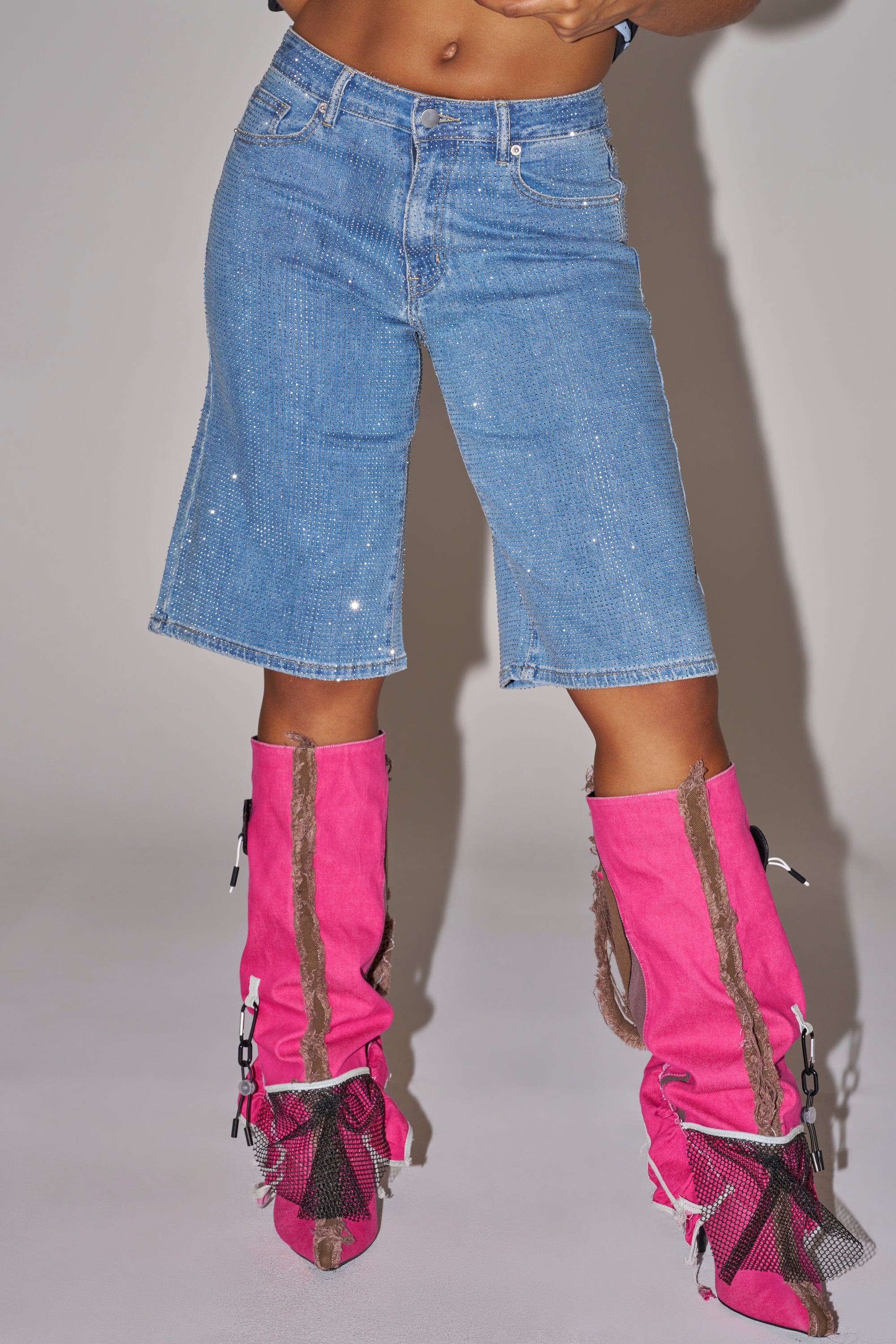 Wearing BLING BLING RHINESTONE DARK BLUE DENIM BERMUDA SHORTS that sparkle, paired with bright pink boots with brown trim and mesh accents, the person stands against a plain light background, upper body not visible. 1
