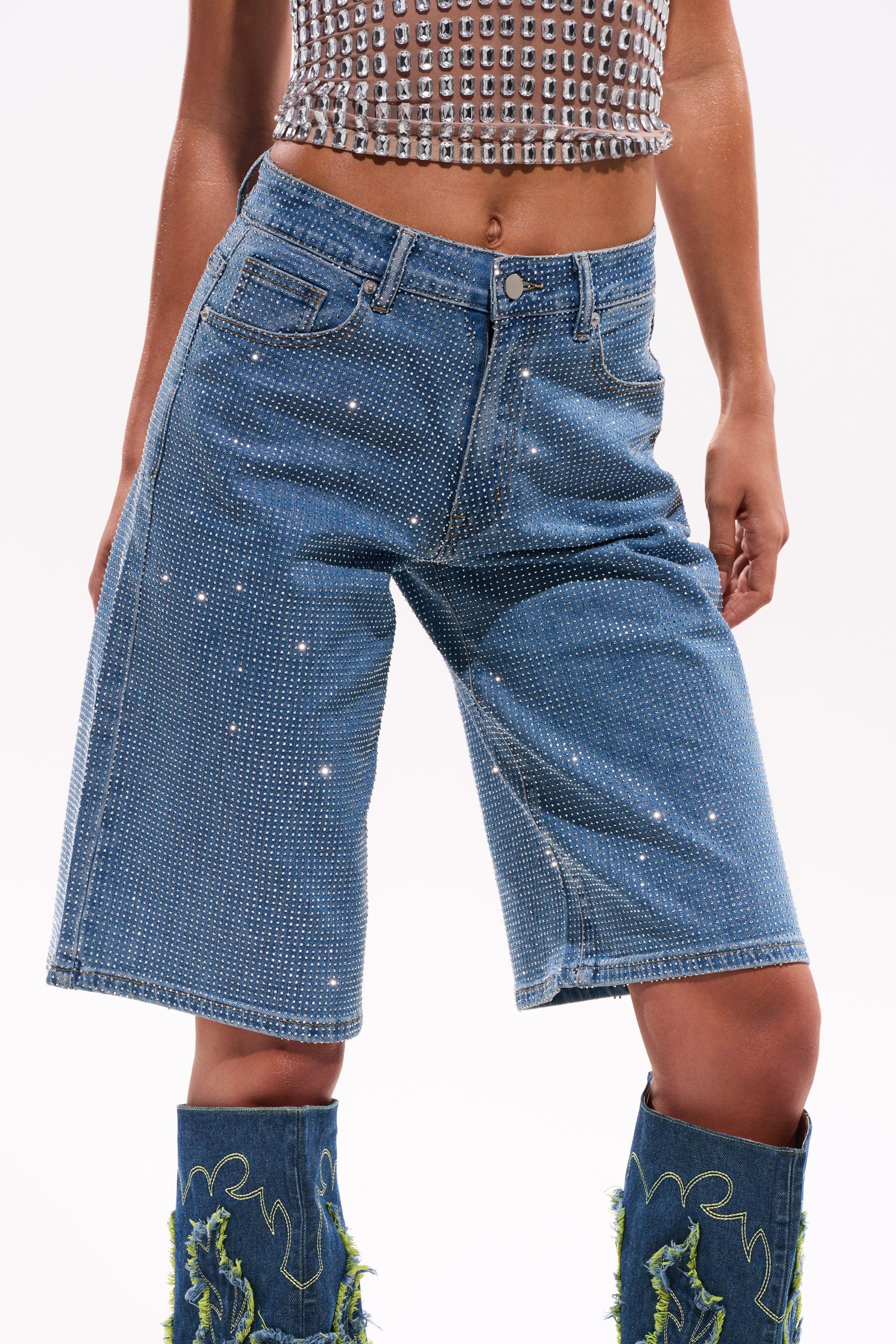 A person wearing the BLING BLING RHINESTONE DARK BLUE DENIM BERMUDA SHORT with a high-rise waist, paired with a cropped studded top and tall blue boots with green fringe, stands against a plain white background. 11
