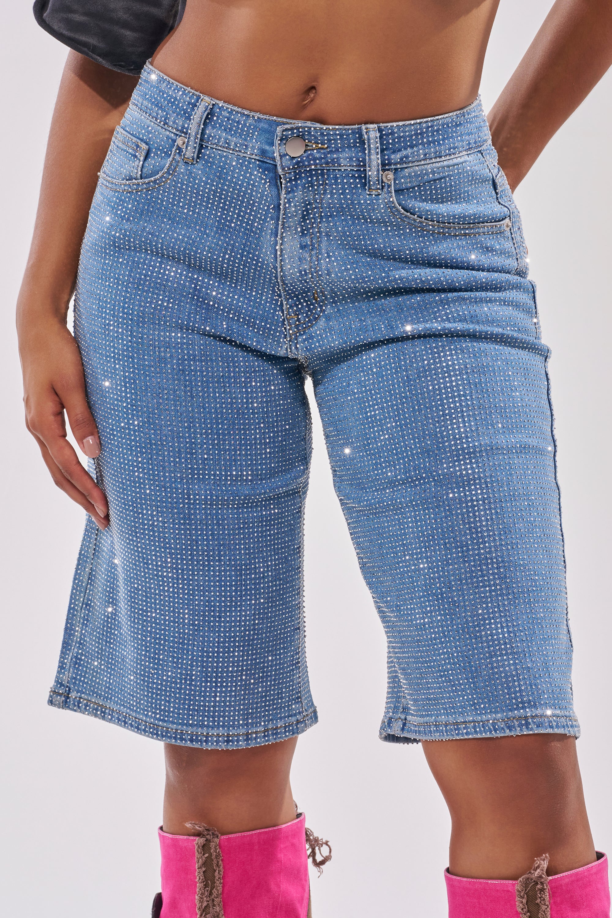 A person wears the BLING BLING RHINESTONE DARK BLUE DENIM BERMUDA SHORT, featuring sparkling blue rhinestones and a fitted, high-rise design, paired with bright pink boots with brown details against a light background. 2