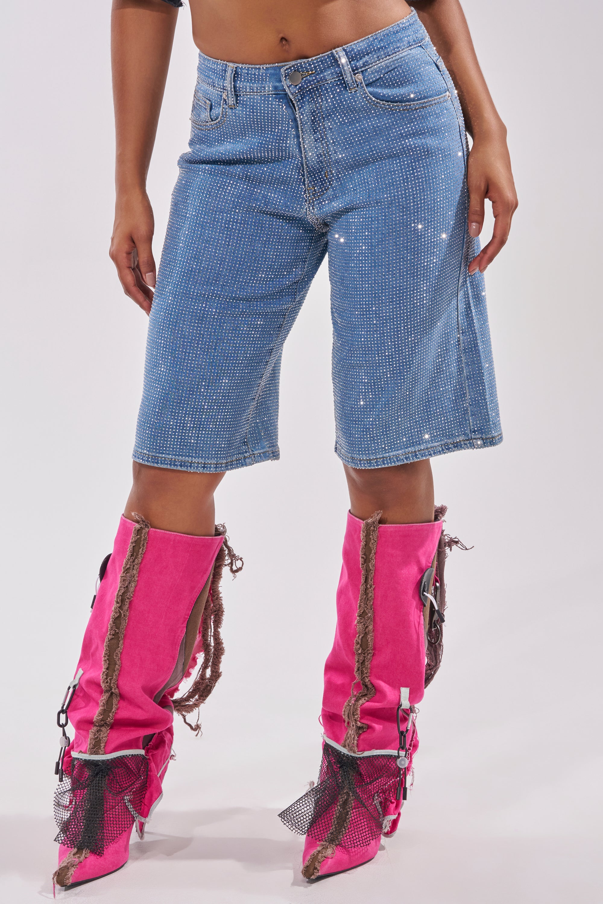 Wearing the BLING BLING RHINESTONE DARK BLUE DENIM BERMUDA SHORTS with bright pink slouchy boots featuring brown rope details and mesh accents, a person stands against a plain light background. 3