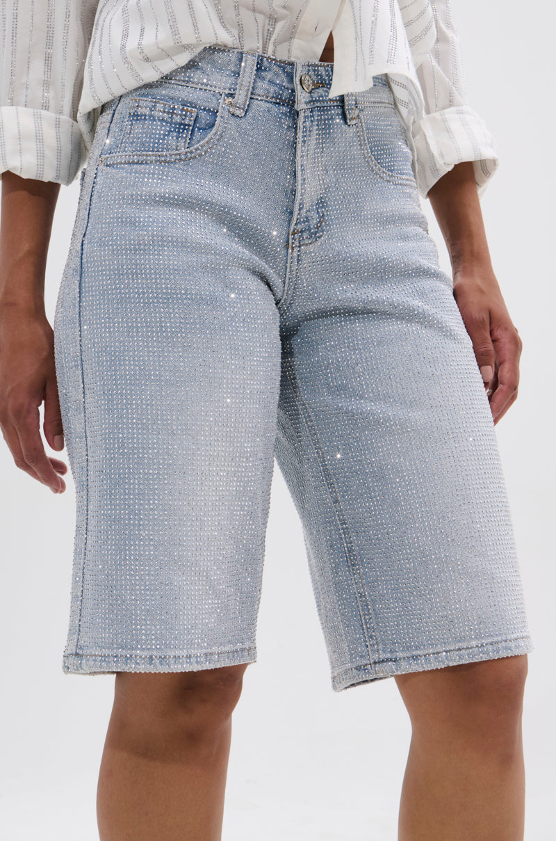 A person wears BLING BLING RHINESTONE BERMUDA SHORT—light-wash, high-rise denim adorned with sparkling rhinestones—paired with a white, long-sleeve, striped button-up shirt.