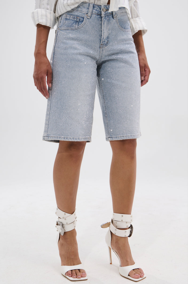 Someone stands against a plain white background wearing the BLING BLING RHINESTONE BERMUDA SHORT in light-wash denim with rhinestone details, paired with a white long-sleeve shirt and white high-heeled buckle-strap sandals.