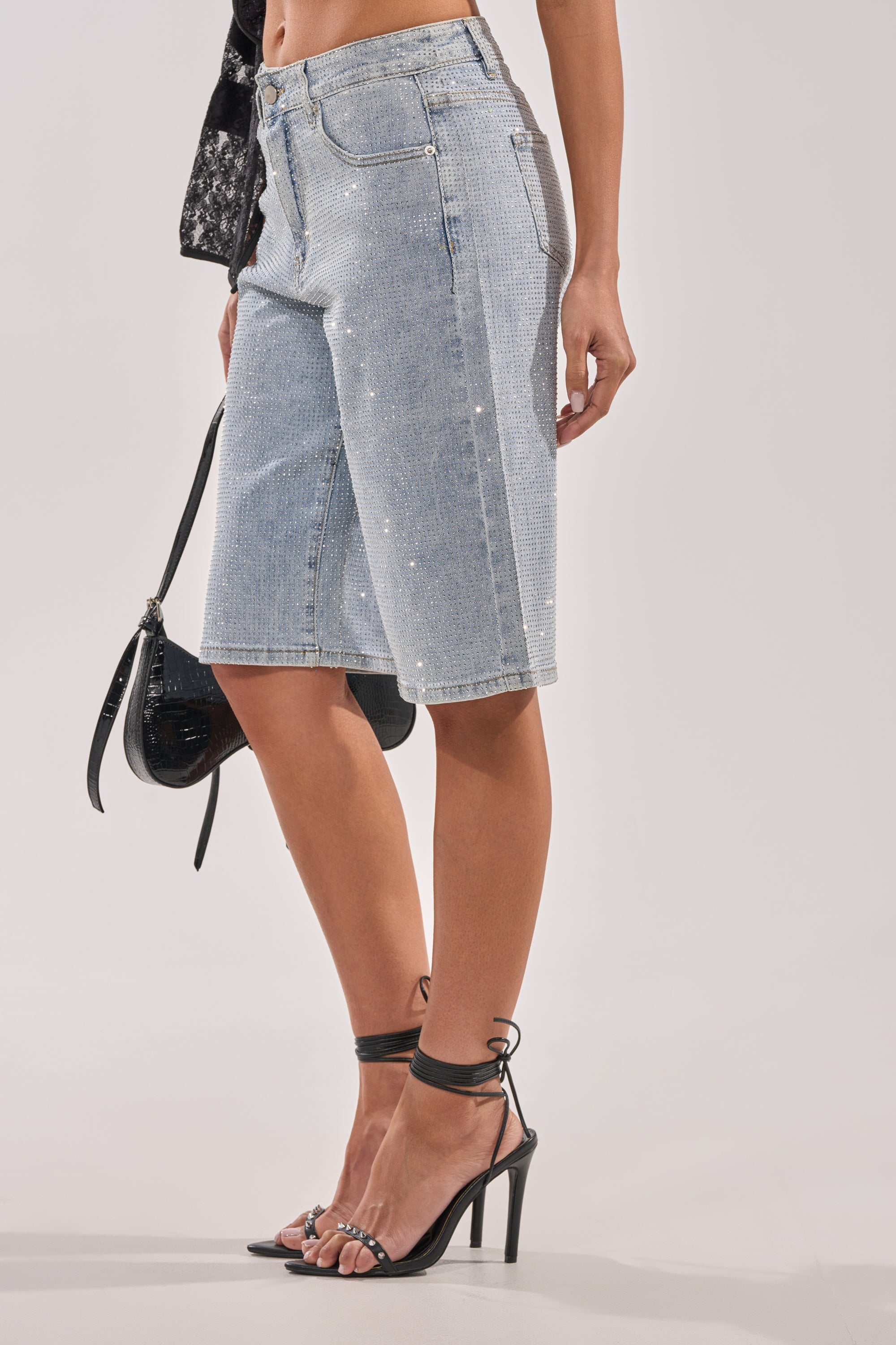 A person stands against a plain background wearing BLING BLING RHINESTONE BERMUDA SHORT IN LIGHT BLUE DENIM, paired with a black handbag and black ankle-strap high-heeled sandals. 2