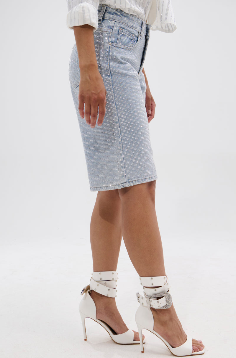 A person wears the BLING BLING RHINESTONE BERMUDA SHORT along with a white long-sleeve top and white ankle-strap high heels, standing against a plain white background.