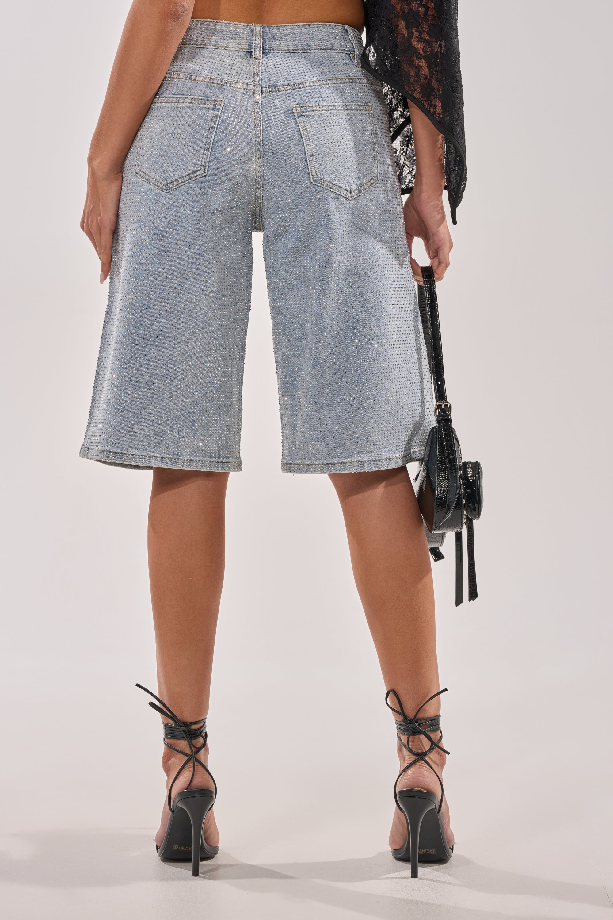 Someone wearing the BLING BLING RHINESTONE BERMUDA SHORT IN LIGHT BLUE DENIM, styled with black high-heeled sandals and a small black handbag, is photographed from behind against a plain background. 3