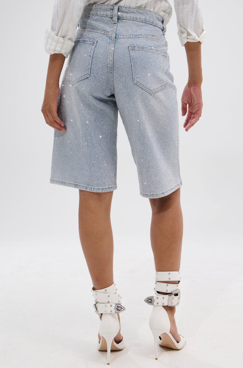 A person stands facing away, wearing the BLING BLING RHINESTONE BERMUDA SHORT in sparkling light-wash denim, paired with a white rolled-sleeve shirt and white high-heeled sandals with multiple straps and silver buckles.