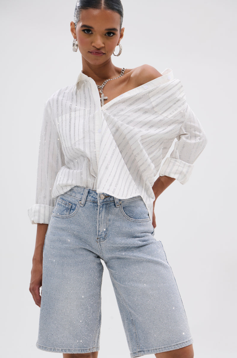 A person poses confidently in a white pinstriped button-up shirt with one shoulder exposed and the BLING BLING RHINESTONE BERMUDA SHORT, accessorized with a necklace and earrings against a plain white background.