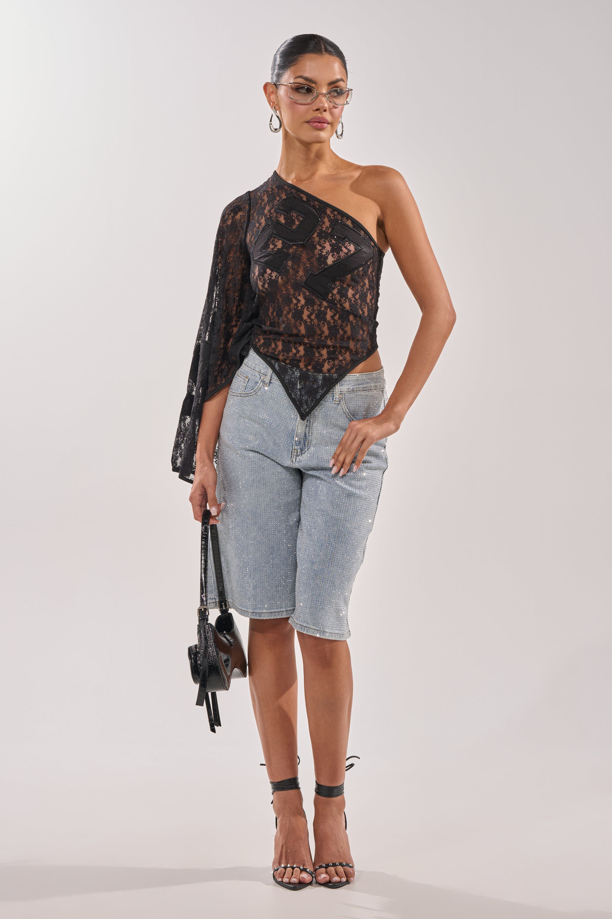 A woman wears the BLING BLING RHINESTONE BERMUDA SHORT IN LIGHT BLUE DENIM with a sheer black lace one-shoulder top, strappy heels, hoop earrings, sunglasses, and holds a small black handbag against a plain white background. 5