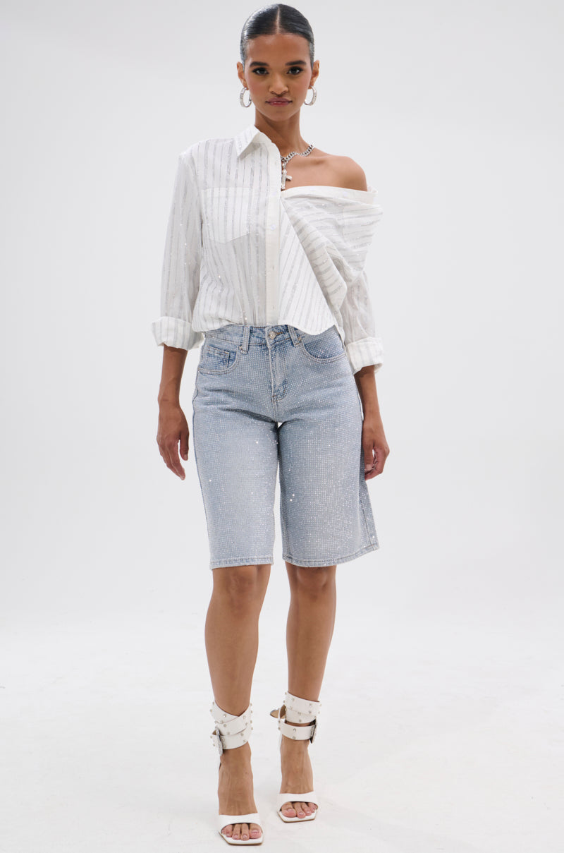 A woman faces forward wearing a partially off-shoulder white striped shirt, the BLING BLING RHINESTONE BERMUDA SHORT in light-wash denim, white ankle-strap heels, and statement jewelry against a plain white background.