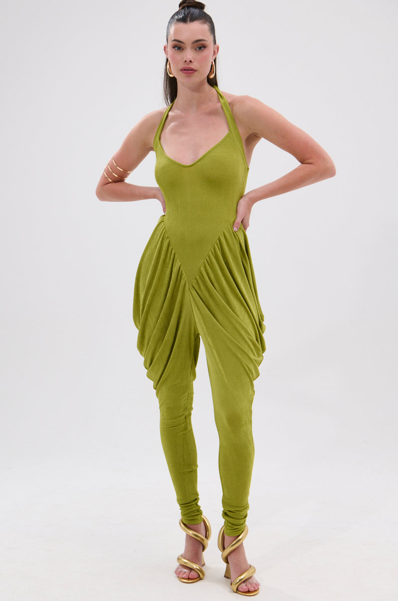 A woman models the UP YOUR GAME RUCHED JUMPSUIT with a halter neckline, draped sides, and tapered legs. She styles it with strappy yellow heels, a gold arm cuff, and a sleek high ponytail. 0
