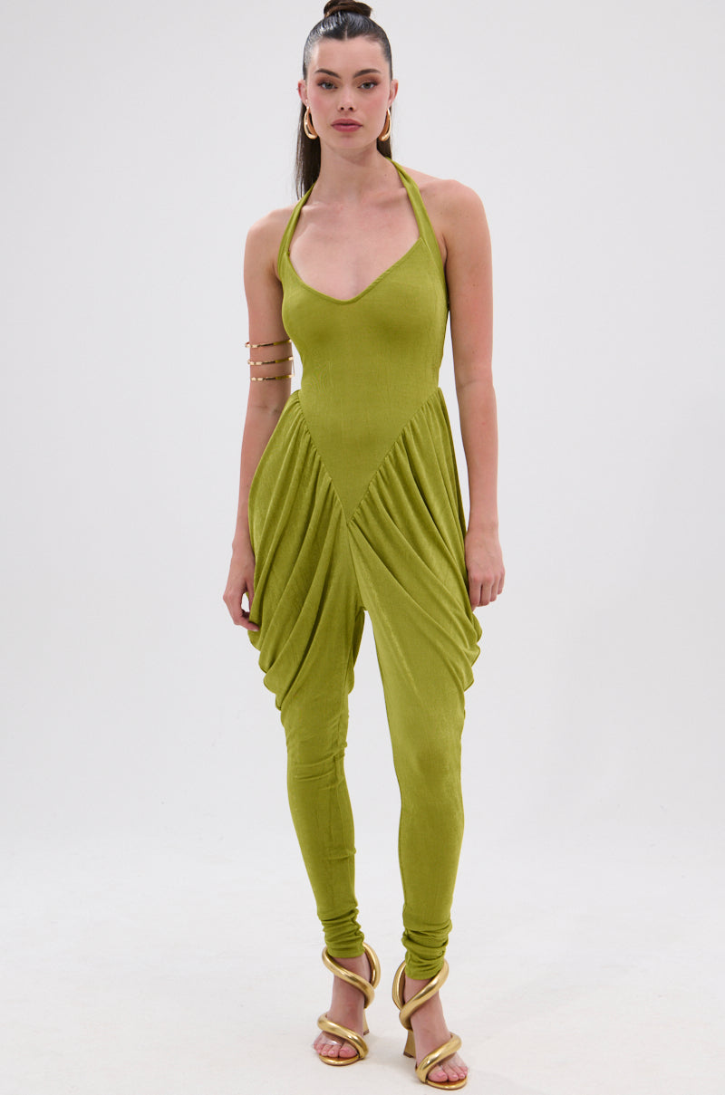 A woman models the UP YOUR GAME RUCHED JUMPSUIT, featuring a sleeveless halter neckline, ruched fabric, and draped hip details. She accessorizes with hoop earrings, gold arm cuffs, yellow strappy heels, and wears her hair in a high ponytail. 1