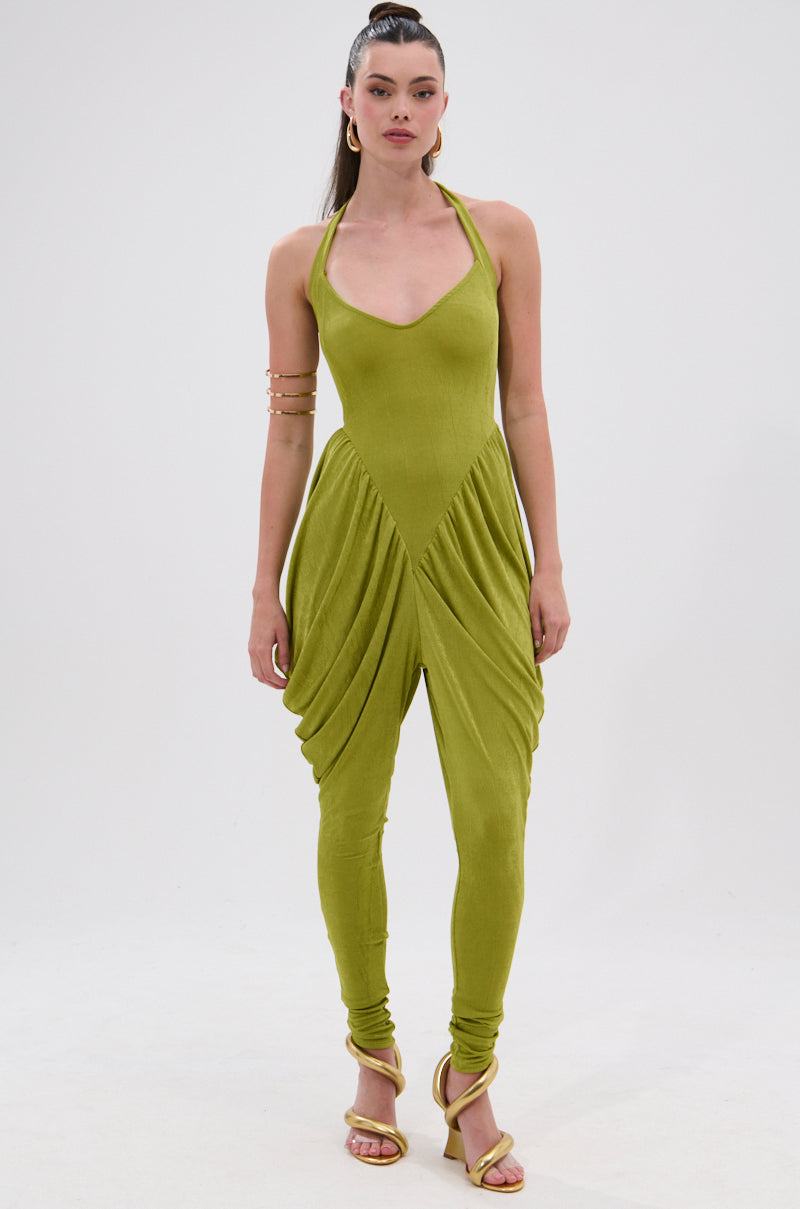A person poses against a plain white background wearing the UP YOUR GAME RUCHED JUMPSUIT in light green, styled with matching heeled sandals, a high ponytail, and gold arm cuffs. Draped detailing and fitted ruching complete the look. 2