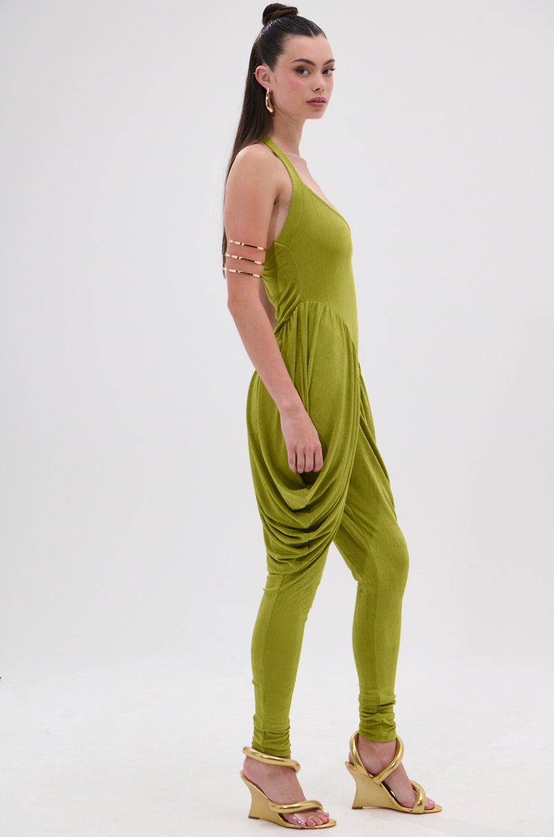 A woman with long dark hair in a high ponytail models the UP YOUR GAME RUCHED JUMPSUIT, a sleeveless olive green wide-leg style, paired with gold wedge sandals, gold arm cuffs, and hoop earrings against a plain white background. 3