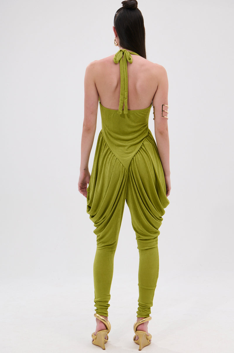 A person faces away, wearing the UP YOUR GAME RUCHED JUMPSUIT in olive green with a halter neckline, draped fabric, and fitted legs. They pair it with heeled sandals, a high bun hairstyle, and a gold upper arm cuff. 4