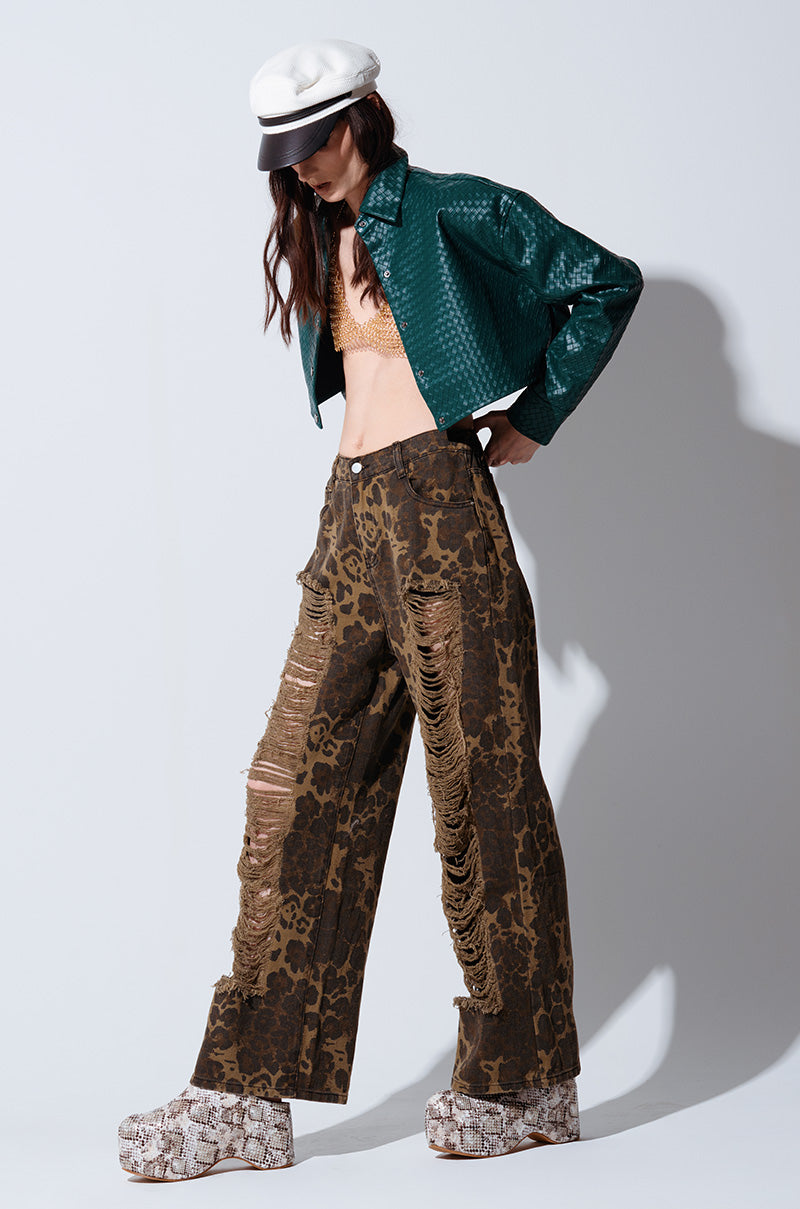 A person with long, dark hair is wearing a white cap, a textured green jacket over a beige bralette, and distressed pants known as the MIMI CHEETAH PANT. They are also sporting platform shoes with a snakeskin pattern. They are posing against a plain light background. 1