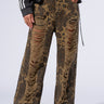 A person is wearing a black and white jacket paired with the MIMI CHEETAH PANT, featuring a brown cheetah print design with mid-rise waist, significant rips, and fraying on the front thighs. They are standing on a light background, which highlights their black platform shoes.