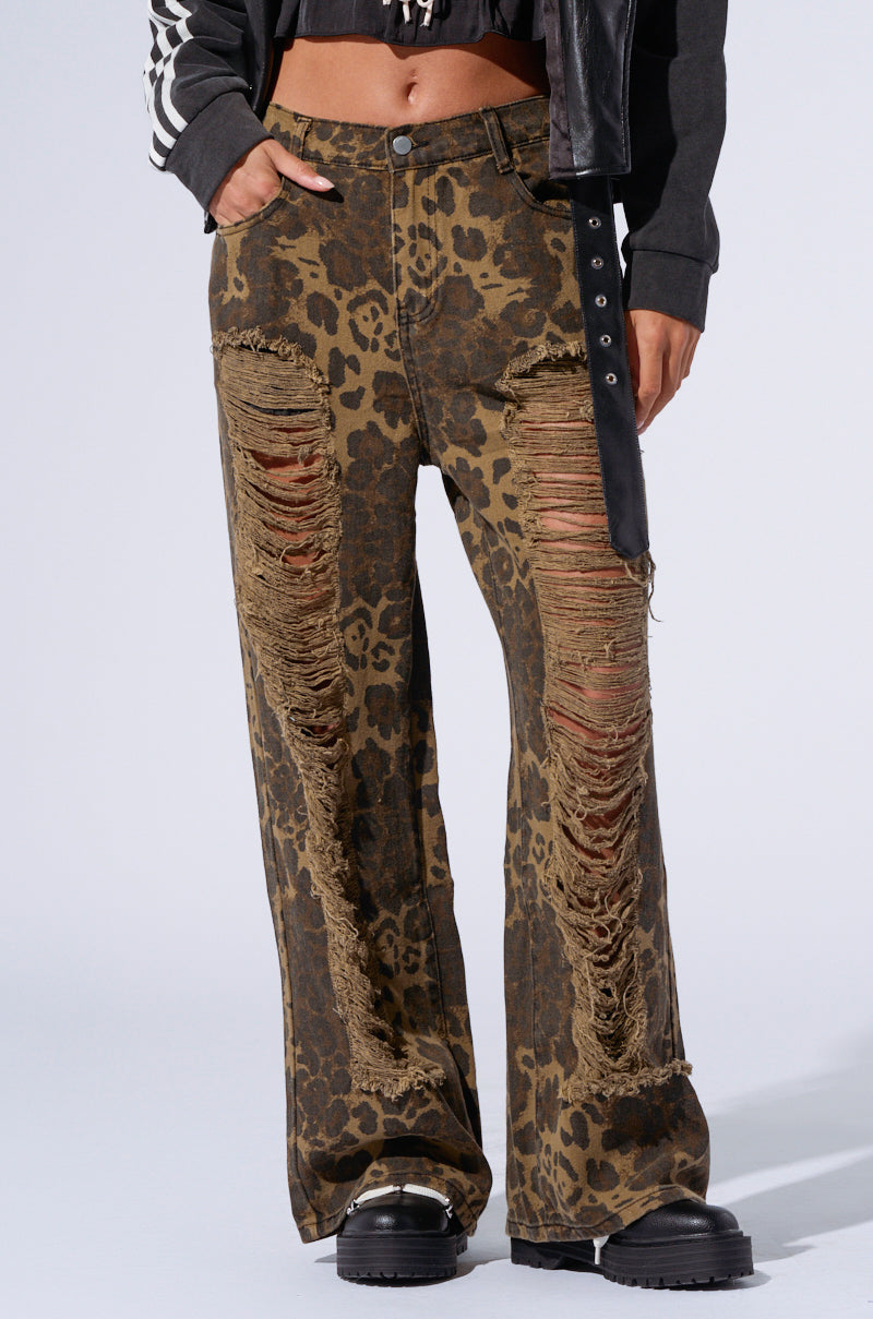 Wearing the striking MIMI CHEETAH PANT, featuring a high-waisted, flared design with a distressed cheetah print, an individual complements the look with chunky black platform boots and a dark top. The pants are notably adorned with prominent rips down the front that reveal light brown fabric underneath. 2