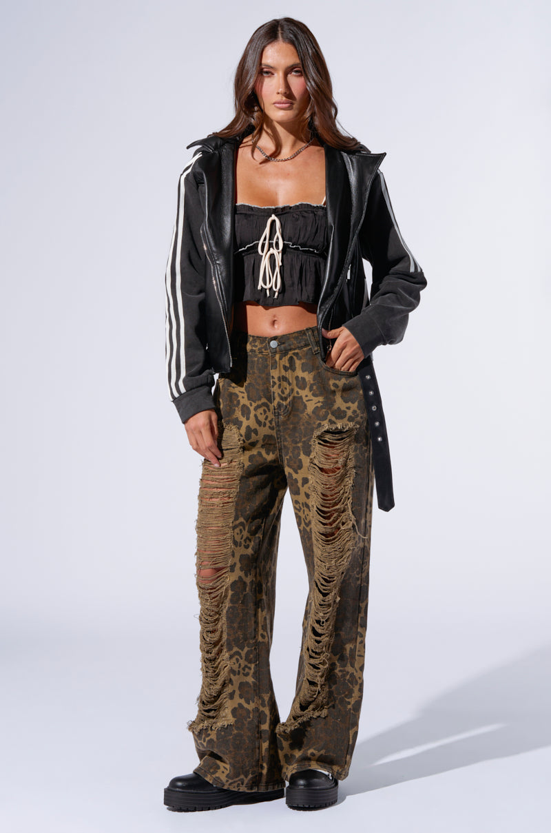 A person stands against a plain backdrop wearing a black leather jacket with white stripes on the sleeves, a black ruched crop top with string detail, and mid-rise MIMI CHEETAH PANT with extensive rips. They also have black chunky platform shoes. 6