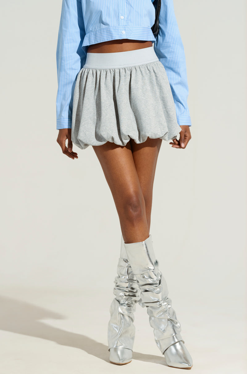 A model showcases a long-sleeve blue top paired with the trendy "LOOK AT ME BUBBLE SKIRT" and metallic silver knee-high boots, posed against a plain backdrop. The focus is on the outfit's chic allure.