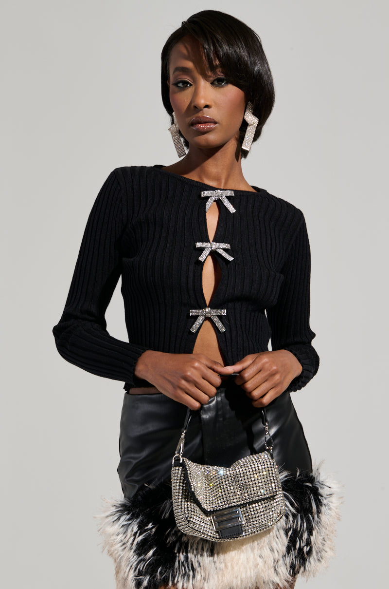 A woman poses confidently in the BEST ONE YET TOP, a stylish black ribbed knit sweater adorned with peekaboo bow cutouts and rhinestone embellishments. She holds a fashionable handbag with sparkling details and fur accents, complemented by large glittering earrings and black leather pants, exuding elegance and style.