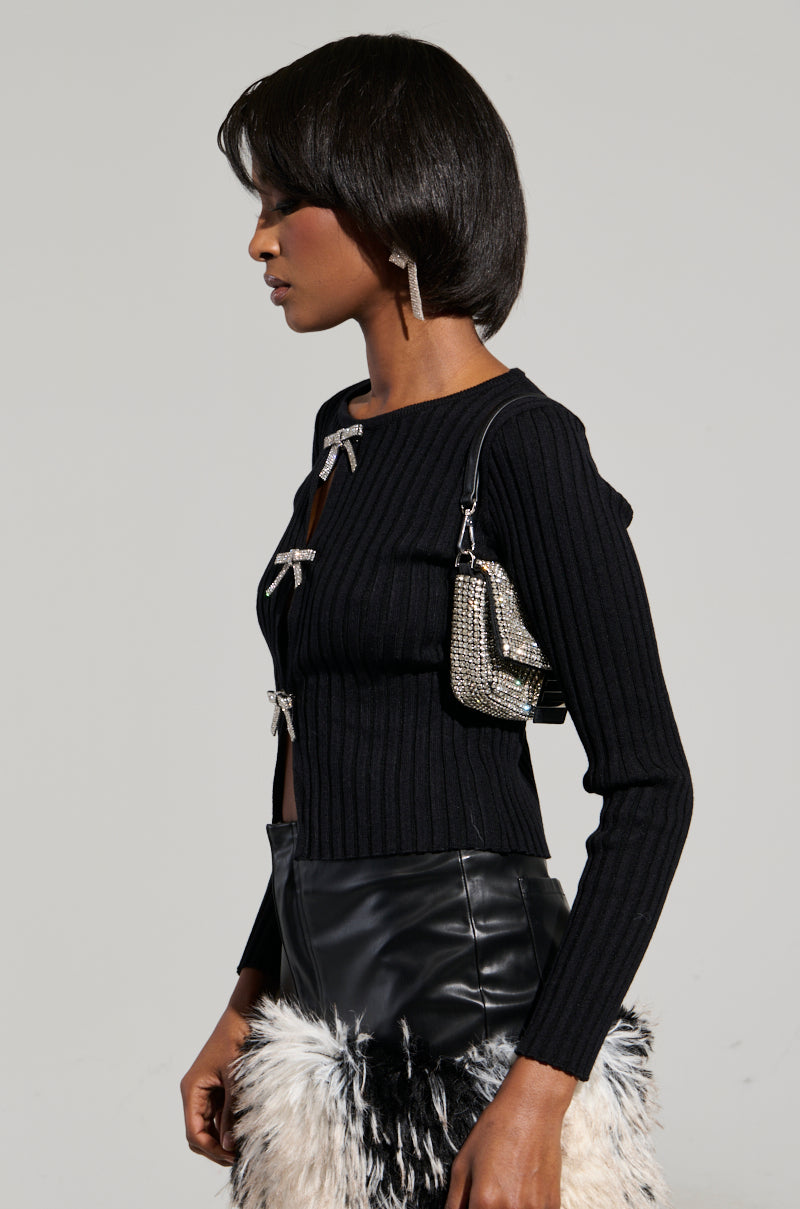 A woman in profile wears the "BEST ONE YET TOP," a black ribbed sweater adorned with bows and peekaboo cutouts. It is paired with a black leather skirt featuring a fluffy white and black section at the bottom, along with a small silver purse. Her short hairstyle complements her long, dangling earrings.