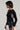 A woman in profile wears the "BEST ONE YET TOP," a black ribbed sweater adorned with bows and peekaboo cutouts. It is paired with a black leather skirt featuring a fluffy white and black section at the bottom, along with a small silver purse. Her short hairstyle complements her long, dangling earrings.