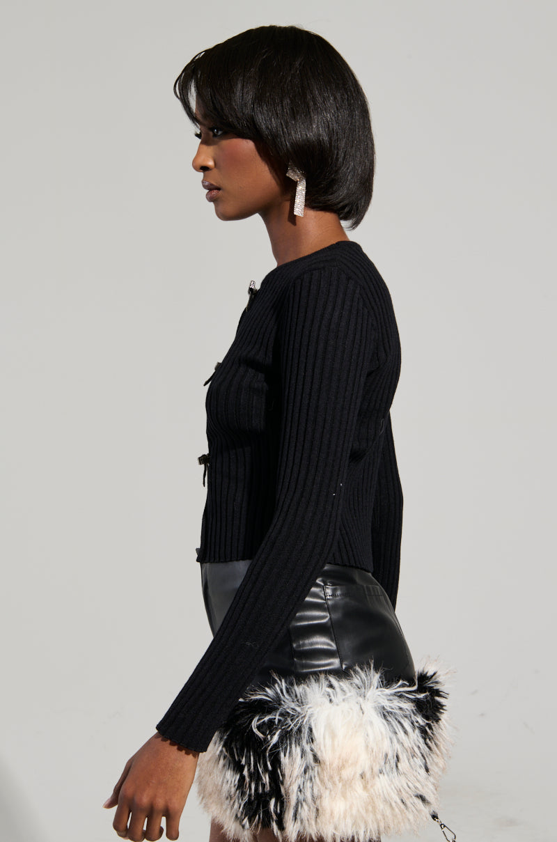 A woman stands in profile against a neutral background, wearing the BEST ONE YET TOP - a ribbed knit black sweater - paired with a leather skirt featuring a fluffy black and white hem. Her short, styled hair complements her rhinestone-embellished, square-shaped earrings.