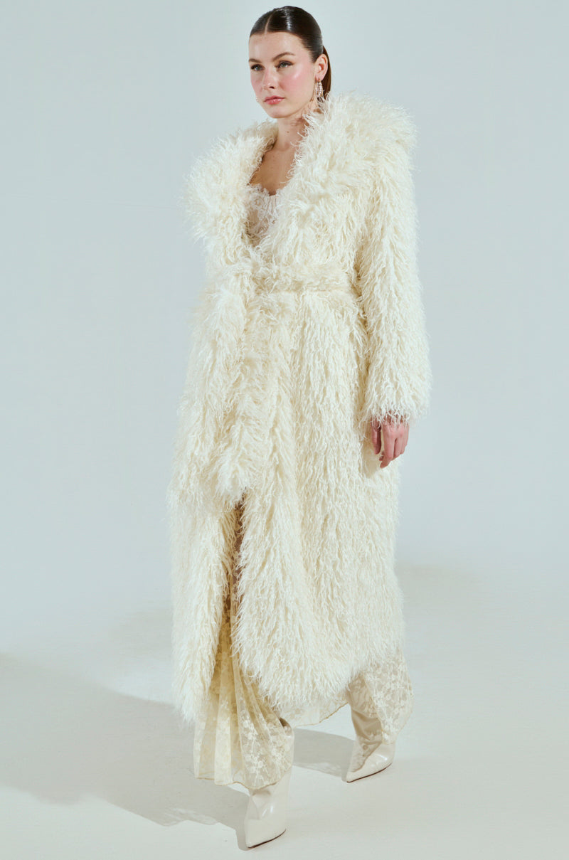 Dressed in the SNOW WHITE TRENCH featuring textured faux fur over a patterned dress, a person stands against a plain backdrop. White platform shoes and a sleek hairstyle complete the ensemble, exuding elegance and modern style.
