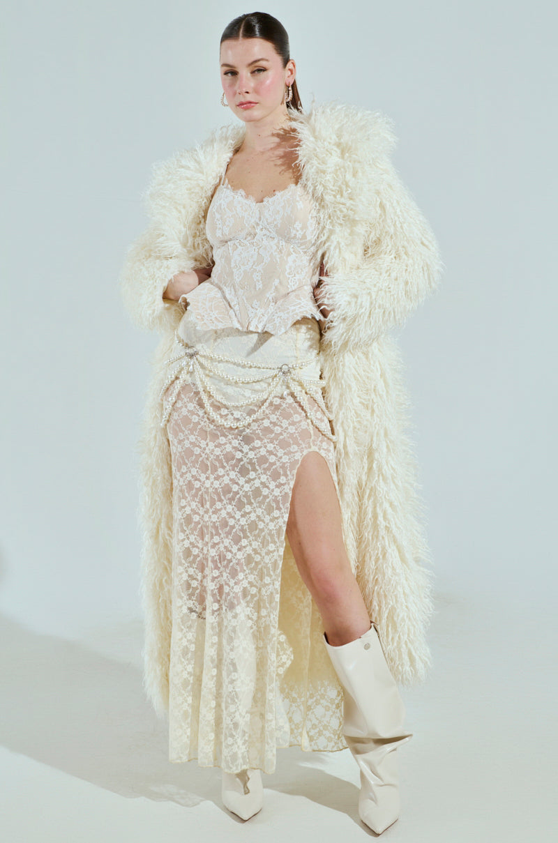 Dressed in a SNOW WHITE TRENCH, a person stands confidently, the textured coat elegantly layered over lace attire. The ensemble includes a high-slit skirt with sleek white boots, set against a neutral backdrop.