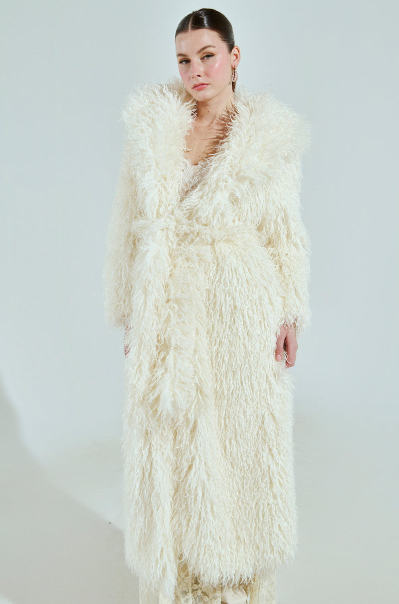 A person wearing the SNOW WHITE TRENCH, a fluffy white faux fur coat, stands against a plain white background. This luxurious belted coat has pockets where they comfortably tuck their hands.