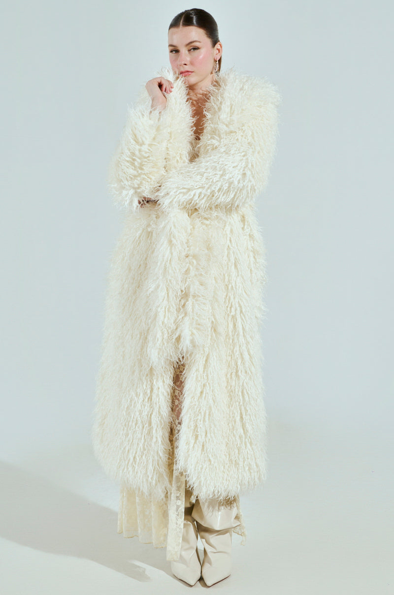 A person poses against a neutral background in a SNOW WHITE TRENCH, characterized by its shaggy, faux fur texture. Their hair is slicked back and they complete the look with light-colored boots.