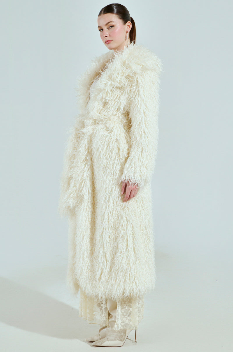A model with long hair in a sleek ponytail showcases the SNOW WHITE TRENCH, a white textured faux fur coat with a waist tie, paired with high-heeled boots against a simple backdrop.
