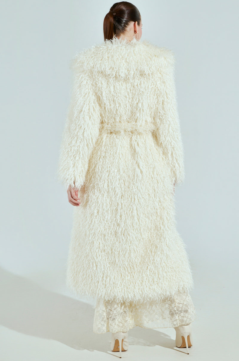 With their brown hair tied back, the person is wearing a long, textured faux fur coat with a belt, sheer lace pants, and cream high heels. The elegant SNOW WHITE TRENCH complements the minimalist scene's plain background.