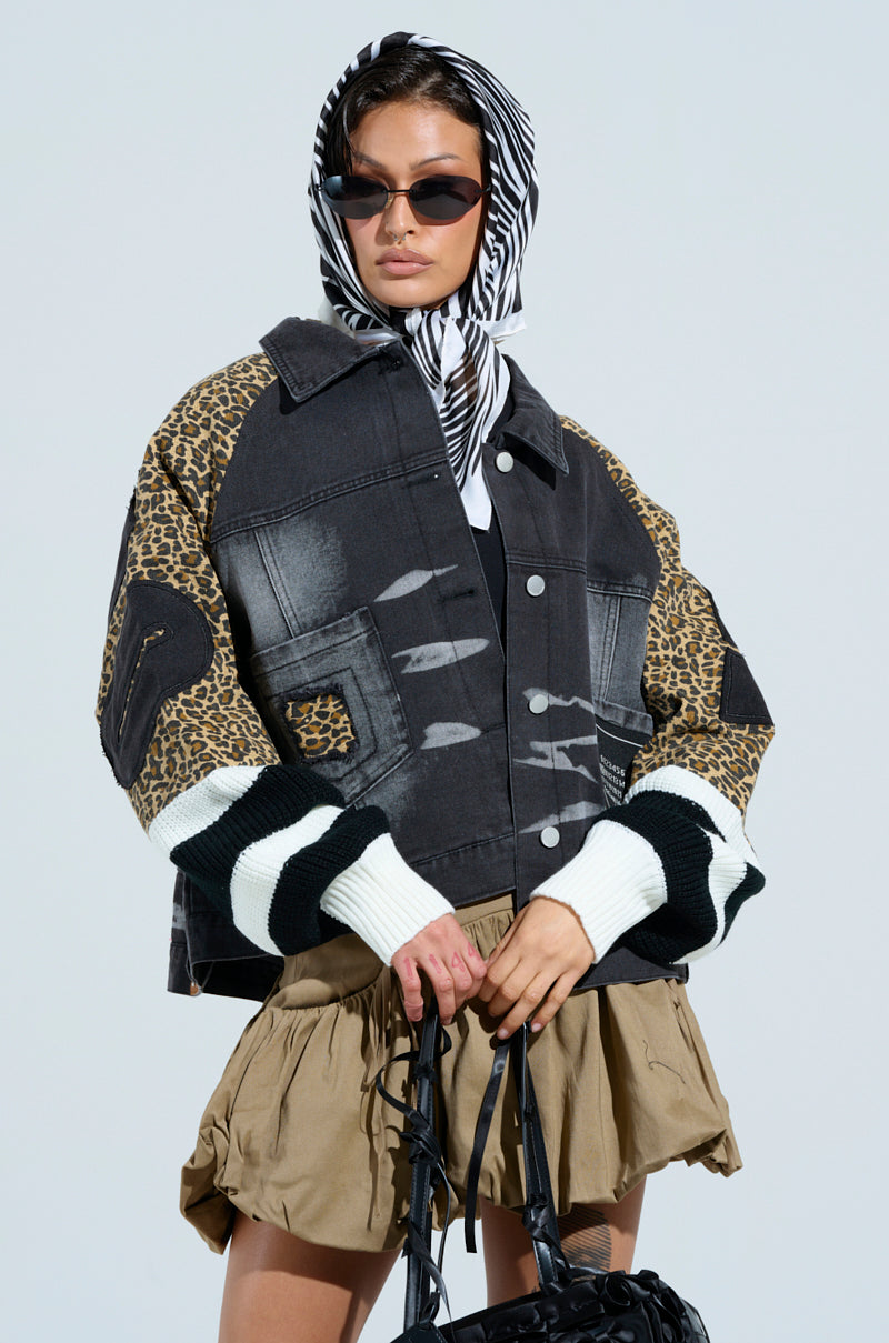 A woman wearing sunglasses and a black-and-white striped headscarf poses confidently. She's styled in a FIERCE CHEETAH BOMBER JACKET, effortlessly paired with distressed denim, and holds a chic black handbag.