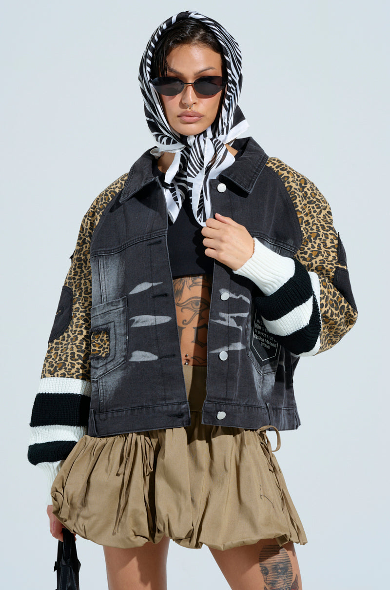 A person wearing the FIERCE CHEETAH BOMBER JACKET, a black crop top, and a tan skirt. They sport a headscarf, sunglasses, and hold a black bag against a light background.