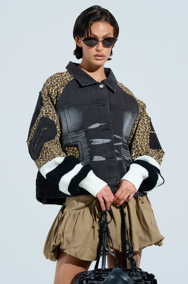 Wearing the FIERCE CHEETAH BOMBER JACKET with its leopard print sleeves and white-black striped cuffs, a person holds a black bag. They're also sporting sunglasses and a khaki skirt, standing against a plain background.