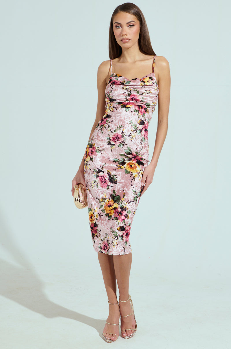 A woman in a CHARLOTTE PRINTED VELVET MIDI DRESS with a cowl neckline poses against a light backdrop, holding a small metallic clutch. She wears strappy heels and her long, sleek hair enhances her confident expression.