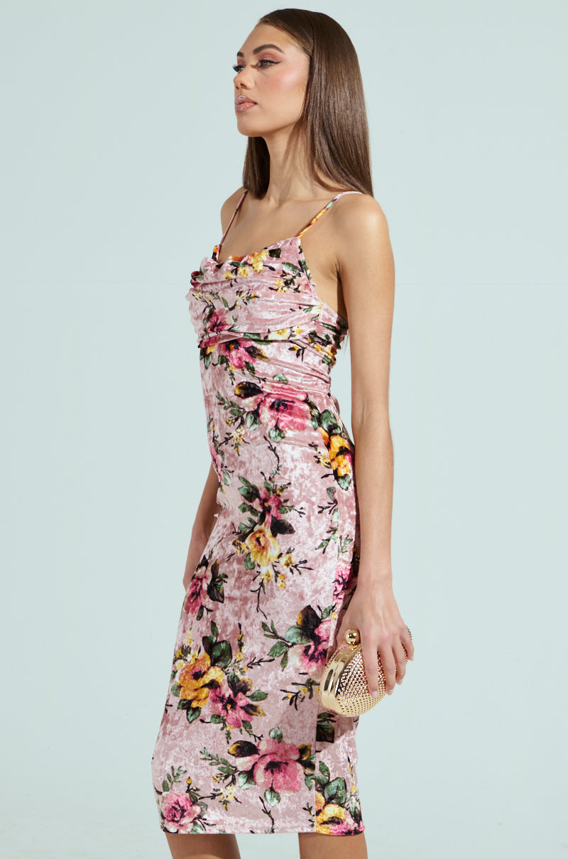 A woman in a CHARLOTTE PRINTED VELVET MIDI DRESS stands sideways, holding a small woven clutch with long, straight brown hair against a light green background.