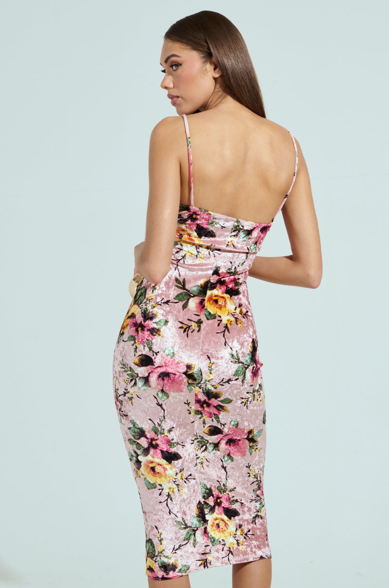 A woman with long brown hair is wearing the CHARLOTTE PRINTED VELVET MIDI DRESS, a sleeveless, form-fitting dress with a pink floral print and thin straps that fall below the knees. She's slightly turned to the side against a plain light blue background.