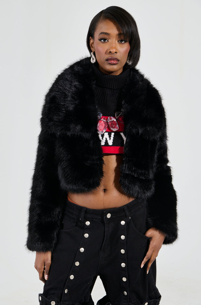 A person wearing the CADENCE FAUX FUR IN BLACK jacket over a cropped sweater featuring a red and white design. They have black pants adorned with button embellishments and are standing against a neutral background.